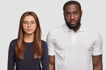 Brother and sister of different races look seriously at camera, stand shoulder to shoulder, listen necessary information, pose for making family portrait isolated on white background. Diverse friends