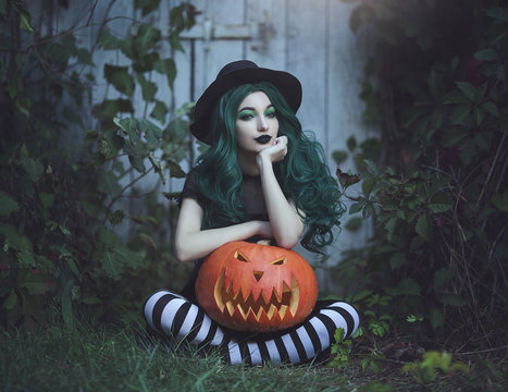Halloween witch with green hair holding a carved scary pumpkin near a wooden door. Beautiful young in a black hat, striped leggings and a suit with a pumpkin. Halloween party art design