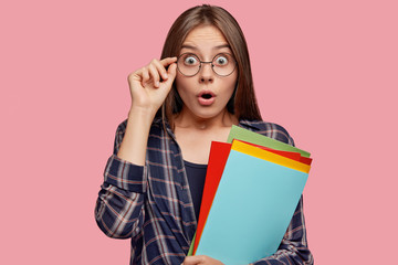 Photo of surprised Caucasian woman stares with bugged eyes, gazes through glasses, carries textbooks, dressed in checkered shirt isolated on pink background. Amazed student has deadline for doing task