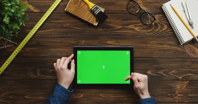 View from above on the black horizontal tablet device on the wooden desk with craftsman equipment, Caucasian female hands scrolling and tapping on the green screen. Chroma key. Tracking motion.