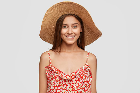 Horizontal Shot Of Comely Girlfriend With Satisfied Expression, Bites Lower Lip, Recieves Compliment From Stranger, Wears Summer Dress, Poses Against White Background. Positive Emotions Concept