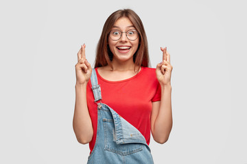 Horizontal shot of pleased smiling teenager keeps fingers crossed, believes in good fortune before...