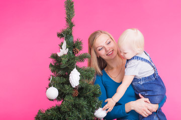 Holidays, family and christmas concept - young woman with her baby near christmas tree on pink background