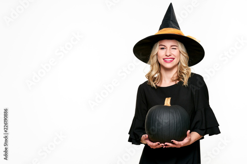 Halloween Witch holding large black pumpkin winking. Beautiful young woman in witches hat and costume holding pumpkin over white background.