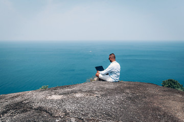 young guy work on laptop on breach .  beautiful sea view. young businessman in sunglasses blue shirt and shorts working with laptop on the rocks