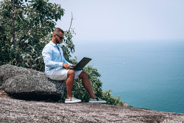 young guy work on laptop on breach .  beautiful sea view. young businessman in sunglasses blue shirt and shorts working with laptop on the rocks