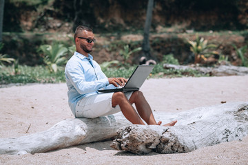 Obraz premium young businessman in sunglasses blue shirt and shorts working with laptop on the beach