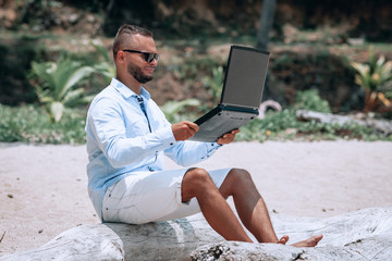 young businessman in sunglasses blue shirt and shorts working with laptop on the beach
