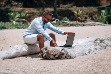 young businessman in sunglasses blue shirt and shorts working with laptop on the beach