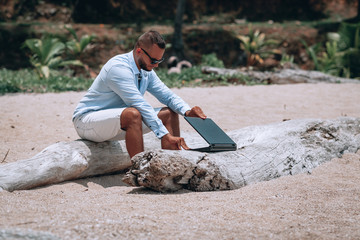 young businessman in sunglasses blue shirt and shorts working with laptop on the beach