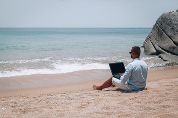 young businessman in sunglasses blue shirt and shorts working with laptop on the beach