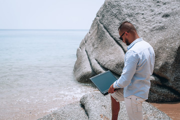 young businessman in sunglasses blue shirt and shorts working with laptop on the beach