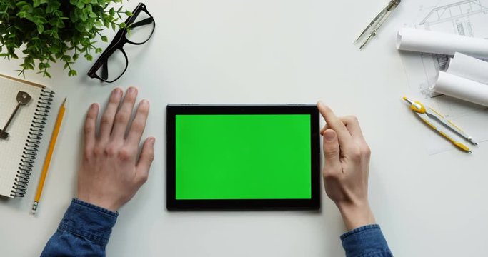 Top view on the male hands scrolling and tapping on the green screen of the black horizontal tablet computer on the designer or architect table. Chroma key.