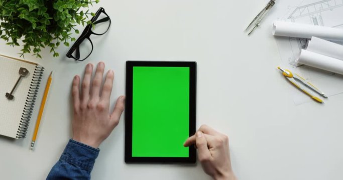 Top view on the male hands scrolling and tapping on the green screen of the black vertical tablet computer on the designer or architect table. Chroma key.