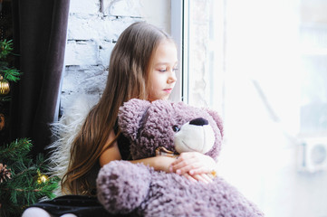 Cute little girl sitting by the window with her Teddy bear and laughing. Girl in a festive dress, dreaming