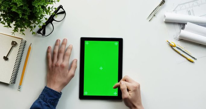Top view on the male hands scrolling and tapping on the green screen of the black vertical tablet computer on the designer or architect table. Chroma key. Tracking motion.