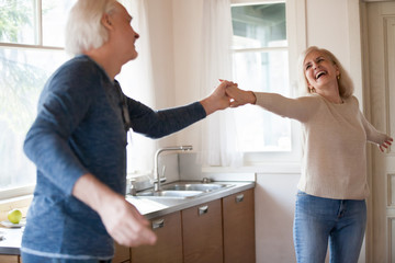 Excited senior woman dancing with mature loving husband in kitchen, old married healthy couple laughing holding hands having fun cooking, active happy middle aged elderly family at home lifestyle