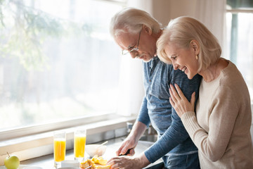 Caring senior husband preparing healthy fresh morning breakfast for happy grateful smiling aged wife laughing embracing loving man in the kitchen, old elderly family couple enjoying cooking together