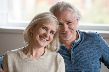 Headshot portrait of happy middle aged romantic couple dating posing indoors, smiling retired old family embracing looking at camera, loving senior mature man and woman hugging bonding together