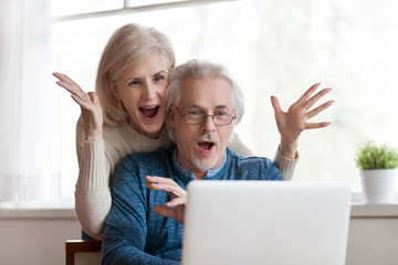 Excited senior mature couple surprised by good unbelievable news, unexpected win, huge shopping sale offer on website, astonished older middle aged family looking at computer screen feeling amazed