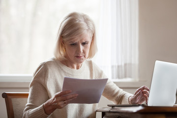 Serious frustrated middle aged senior woman worried reading bad news in paper letter document,...