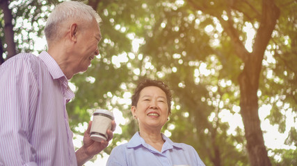 Happy Asian elderly couple morning walk in green city while drinking coffee and talking