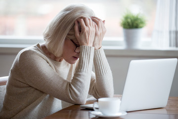 Upset depressed mature middle aged woman in panic holding head in hands in front of laptop...