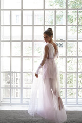 Rear full length view of gorgeous beautiful young European female posing at large window in puffy long pink dress, looking down with shy facial expression. Elegance, luxury, glamour and style