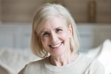 Happy middle aged elderly grey haired woman looking at camera posing at home indoors, positive satisfied single mature senior retired lady with wide toothy smile headshot portrait, natural old beauty