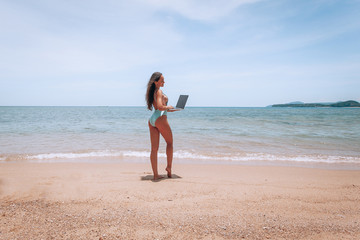 Fototapeta na wymiar Young american woman in blue swimsuit working with laptop on empty beach. Concept of resting on morning sea and summer vacations, modern technology.
