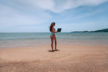 Young american woman in blue swimsuit working with laptop on empty beach. Concept of resting on morning sea and summer vacations, modern technology.