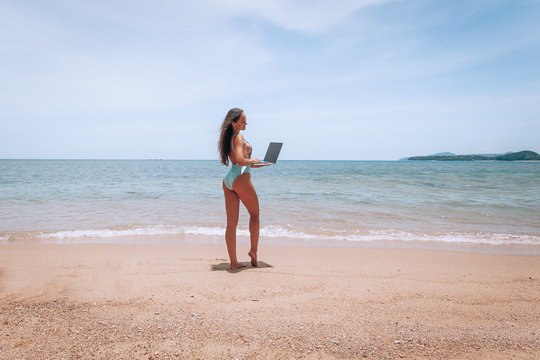 Young american woman in blue swimsuit working with laptop on empty beach. Concept of resting on morning sea and summer vacations, modern technology.