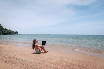 Fototapeta na wymiar Beautiful woman is sitting on the sand, working at the computer with internet , in a white bathing suit, background of sea blue water.
