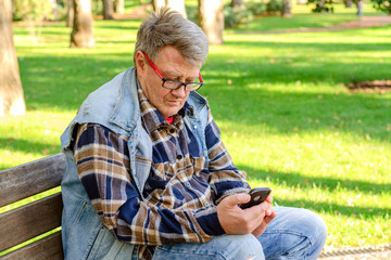 Senior adult man in casual clothing and a eyeglasses, sits on a park bench and enjoys his smartphone. Concept of active leisure for middle-aged and older people.
