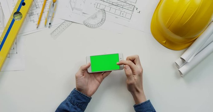 View from above on the male hands scrolling and tapping on the chroma key screen of the white horizontal smartphone on the builder desk with drawings stuff and helmet. Green screen.