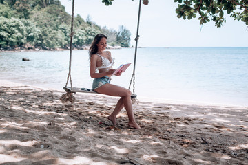 Young fit girl in denim shorts and a white bra swinging on a swing on the shore of the Andaman sea in Phuket. that the writes and reads in Notepad