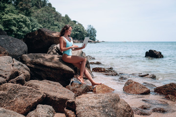 Young happy woman using laptop and sitting on stone near sea, empty morning beach. Concept of modern technology and working on nature, summer vacations.