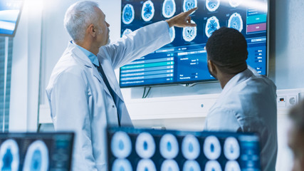 Team of Scientists Work in the Brain Research Laboratory, Discussing Brain Scans Show on Wall TV Monitor. Neurologists / Neuroscientists Surrounded by Monitors Showing CT, MRI Scans.