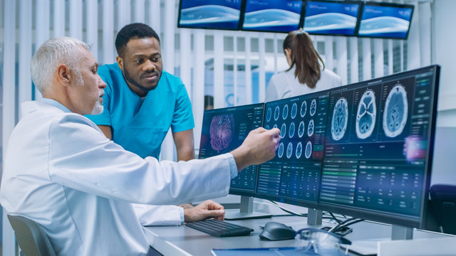 Medical Scientist and Surgeon Discussing CT / MRI Brain Scan Images on a Personal Computer in Laboratory. Neurologists / Neuroscientists in Neurological Research Center Working on a Brain Tumor Cure.