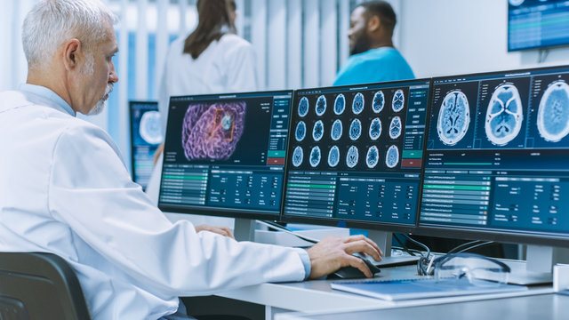 Experienced Senior Scientist Working with CT/ MRI Brain Scan Images on a Personal Computer in Laboratory. Neurologists / Neuroscientists in Medical Research Center Work on Brain Tumor Cure.
