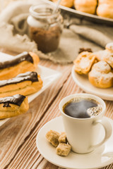 Home cooked profiteroles and eclairs with a cup of black coffee on a wooden table.