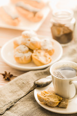 Home cooked profiteroles and eclairs with a cup of black coffee on a wooden table.