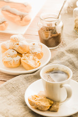 Home cooked profiteroles and eclairs with a cup of black coffee on a wooden table.
