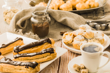 Home cooked profiteroles and eclairs with a cup of black coffee on a wooden table.