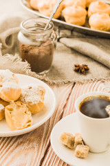 Home cooked profiteroles and eclairs with a cup of black coffee on a wooden table.