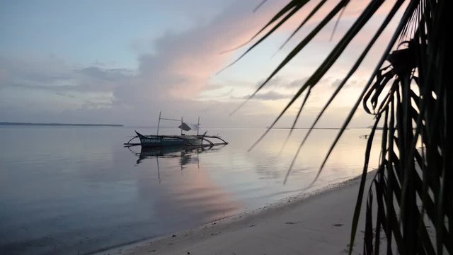 Slow motion shot of traditional filipino bangka boat on calm waters at dusk as seen from beach behind palm tree