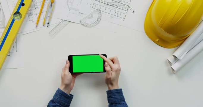 View from above on the female hands scrolling and tapping on the chroma key screen of the black horizontal smartphone on the builder desk with drawings stuff and helmet. Green screen.