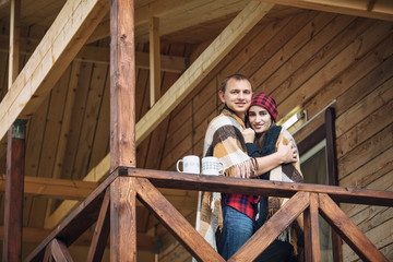 Couple man and woman young beautiful happy on the porch of a wooden house in nature with mugs of hot drinks