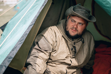 Fototapeta premium Young man in hat, drinking tea or coffee in mountains. Mountain camping concept. Traveler man with beard sit near the tent neat the mountain lake . Man traveler hands holding cup of tea.