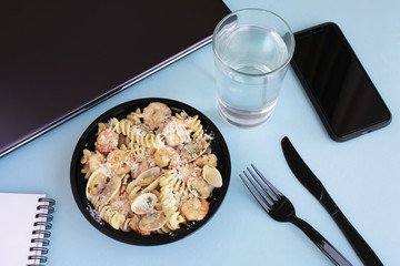 Seafood pasta business lunch at workplace. Open lunch box, closed laptop, knife and fork, smartphone, glass of water, notebook on blue background.
